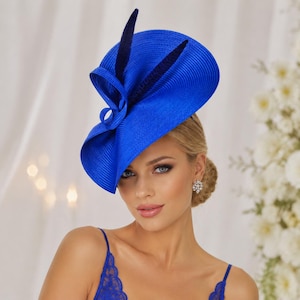 Op de afbeelding: Een levendige koningsblauwe fascinator met een brede, gestructureerde rand met een textuur van ribbels. Het ontwerp bevat een elegant, zwierig lusdetail en twee donkerblauwe veeraccenten die vanuit het midden omhoog steken.