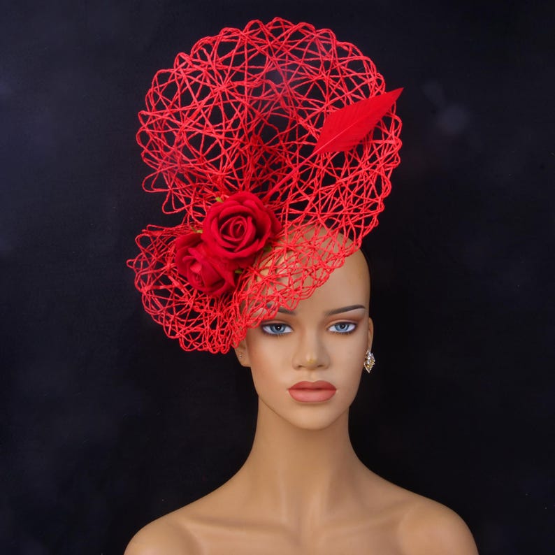 May include: A vibrant red fascinator hat featuring an intricate, web-like wire structure. The headpiece is adorned with two deep red fabric roses and a single matching red feather, creating a bold and sculptural accessory for formal headwear.