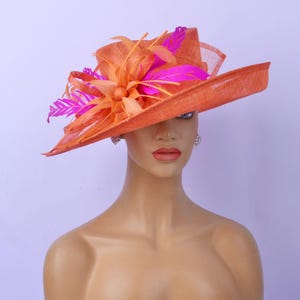 Könnte beinhalten: Ein großer, breitkrempiger Hut in leuchtend orange mit einer rosa und orangefarbenen Blume und Federn an der Seite.