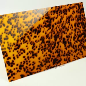 May include: A rectangular sheet of acrylic material featuring a glossy finish with a classic tortoiseshell pattern. The surface displays a repeating organic design of dark brown and black spots layered over a translucent amber and golden yellow base.