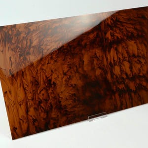 May include: A rectangular panel with a high-gloss finish featuring a swirling, marbled pattern in shades of dark brown, amber, and black. The surface has a reflective quality and is supported by a small, clear acrylic stand at the bottom edge.