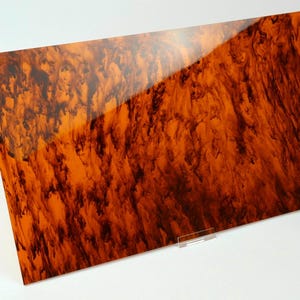 May include: A rectangular sheet of acrylic material featuring a glossy finish with a marbled pattern of deep brown and vibrant orange swirls. The surface is smooth and reflective, displaying a complex, organic design across the entire flat panel.