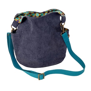May include: A navy blue corduroy shoulder bag with a teal adjustable strap and brass-colored hardware. The bag has two exterior pockets and a patterned interior lining with purple and yellow accents. The bag is designed for everyday use.