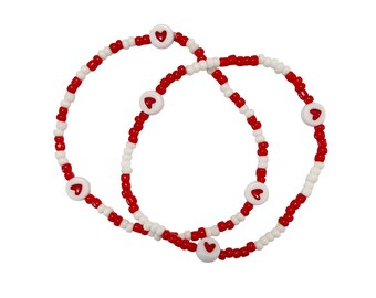 Può includere: Due braccialetti di perline. I braccialetti sono fatti di perline rosse e bianche. Perline bianche con disegni a cuore rosso sono intervallate nei braccialetti. I braccialetti sono su sfondo bianco.