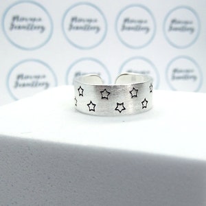 May include: A silver adjustable ring with a matte finish. The ring features small black star cutouts around the band. The ring is presented on a white surface. The ring is likely made of sterling silver.