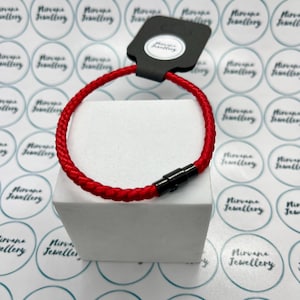 May include: A vibrant red braided bracelet with a black clasp. The bracelet is presented on a white cube, with a black tag attached. The background features a pattern of circular logos.
