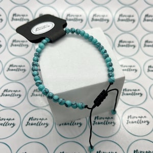 May include: Turquoise beaded bracelet with black detailing. The bracelet is strung on a black cord with adjustable sliding knots. A black tag with the text "Nirvana Jewellery" is attached.