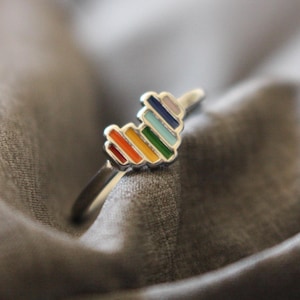May include: A silver ring featuring a rainbow heart design. The heart is made up of small, colourful bars.