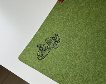 Könnte beinhalten: Eine rechteckige Schreibtischunterlage aus grünem Filz mit einer schwarzen Strichzeichnung von zwei Pilzen. Am oberen Rand befindet sich ein kleines braunes, rechteckiges Etikett. Die Oberfläche hat eine texturierte Optik auf einem weißen Tisch.