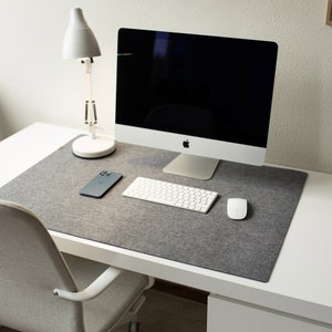 May include: A white desk with a gray felt desk mat, a white lamp, a computer monitor, a keyboard, a mouse, and a smartphone.
