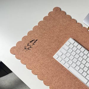 Puede incluir: Un espacio de trabajo inform&aacute;tico con un port&aacute;til gris, un teclado blanco y una alfombrilla de escritorio de fieltro marr&oacute;n con borde festoneado y dise&ntilde;o de ramita de lavanda. El conjunto est&aacute; sobre un escritorio blanco.