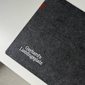 Peut inclure: Un sous-main rectangulaire en feutre gris foncé avec le texte brodé en blanc "Gerhard's Lieblingsplatz" dans le coin inférieur gauche et une petite étiquette rectangulaire brune fixée sur le bord supérieur gauche du feutre.