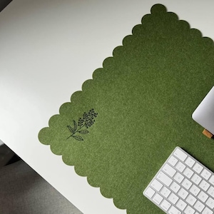 Puede incluir: Una alfombrilla de escritorio rectangular de fieltro verde oliva con borde festoneado y una ilustración botánica de líneas negras de una ramita de flores en la esquina. La alfombrilla sirve de base para un portátil gris sobre un soporte de madera y un teclado inalámbrico blanco.