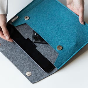 Può includere: Una custodia in feltro color verde acqua e grigio con patta ripiegabile e due chiusure magnetiche circolari argentate. L'interno è foderato in feltro grigio e contiene un tablet nero e uno smartphone nero riposti nello scomparto.