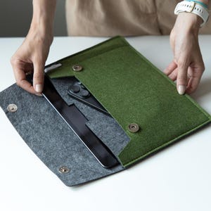 May include: A green felt tablet and phone case with a gray felt interior. The case is open, revealing a black tablet and a black smartphone. The case has a magnetic snap closure and is being held by a person.