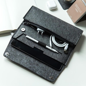 May include: A gray felt organizer case with a zipper closure, snap buttons, and multiple compartments. Inside are a tablet, stylus, charging cables, and a power adapter. The case is open, displaying the contents.