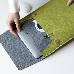 Könnte beinhalten: Eine Laptoptasche aus lindgrünem und anthrazitfarbenem Filz mit einem Umschlag und zwei runden, silbernen Magnetverschlüssen. Im Inneren befinden sich ein silberner Laptop, ein weißes Ladekabel und zwei kleine silberne Metall-Eingabestifte.