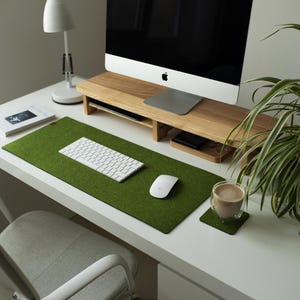 May include: A white desk with a green felt desk mat, a white keyboard, a white mouse, a wooden monitor stand, a cup of coffee on a coaster, and a potted plant.