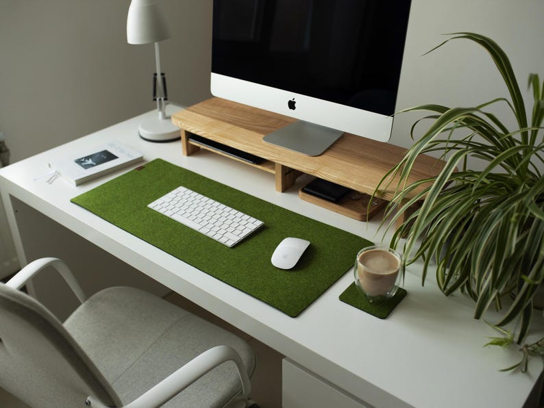 May include: A white desk with a green felt desk mat, a white keyboard, a white mouse, a wooden monitor stand, a cup of coffee on a coaster, and a potted plant.