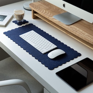 May include: A dark navy blue felt desk mat with a scalloped edge design. The rectangular mat features a soft, textured surface and is positioned under a white wireless computer keyboard and a white computer mouse on a white desk surface.