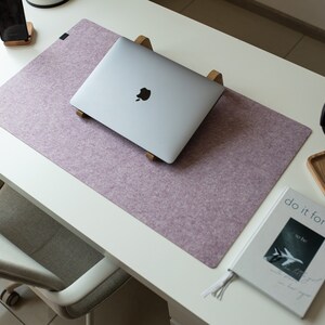 May include: A purple felt desk mat with a laptop computer on a wooden stand, a smartphone on a wooden stand, a book with the text "do it for her" and a pair of black headphones on a wooden surface.