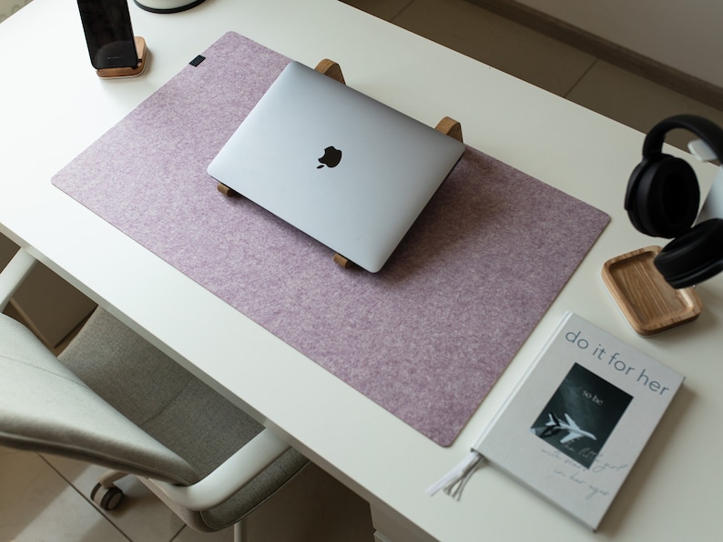May include: A purple felt desk mat with a laptop computer on a wooden stand, a smartphone on a wooden stand, a book with the text "do it for her" and a pair of black headphones on a wooden surface.