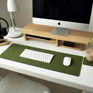 May include: A white desk with a green felt desk mat, a white keyboard, a white mouse, a wooden monitor stand, a white lamp, a black headphone stand, and a glass cup with a brown drink on a brown coaster.