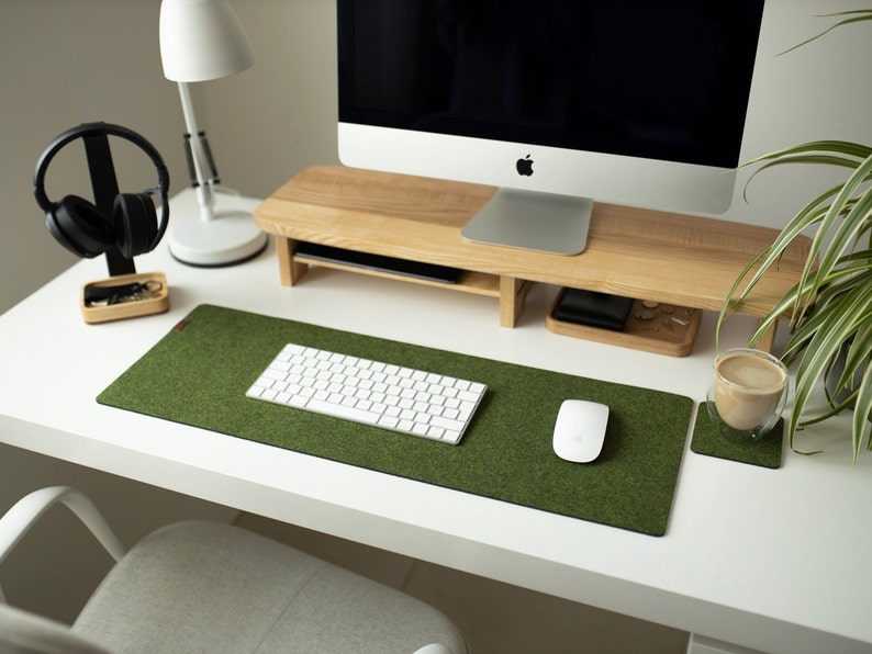 May include: A white desk with a green felt desk mat, a white keyboard, a white mouse, a wooden monitor stand, a white lamp, a black headphone stand, and a glass cup with a brown drink on a brown coaster.
