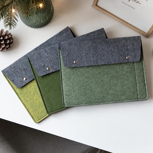 May include: Three rectangular felt sleeves in varying shades of green, each featuring a dark gray fold-over flap secured with a single silver-toned metal snap button. The sleeves are arranged in a layered, overlapping stack on a flat surface.