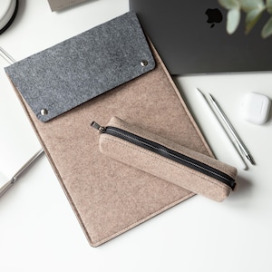May include: A beige felt document folder with a dark gray felt flap secured by two silver metal snaps, accompanied by a matching beige felt pencil case featuring a black zipper closure, positioned alongside a silver pen and white wireless earbuds.