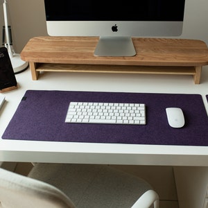 May include: A white desk with a purple felt desk mat, a white wireless keyboard, a white wireless mouse, a wooden monitor stand, a smartphone, a book, and a glass of water on a coaster.