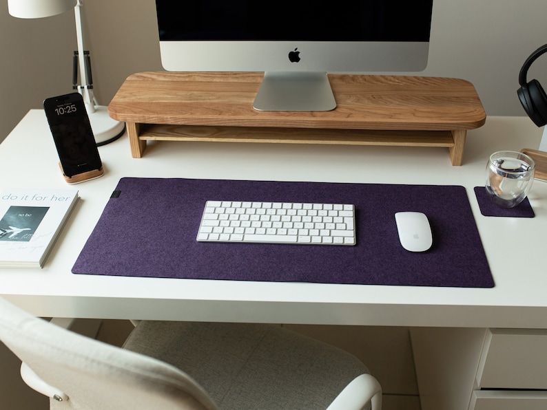 May include: A white desk with a purple felt desk mat, a white wireless keyboard, a white wireless mouse, a wooden monitor stand, a smartphone, a book, and a glass of water on a coaster.