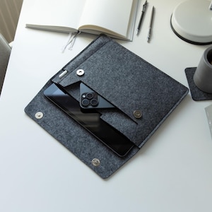 May include: A grey felt tablet and phone case with a magnetic fastening, holding a black tablet and phone. The case is open, revealing the devices inside. A pen is tucked into a side pocket. The scene is set on a white desk with office supplies.
