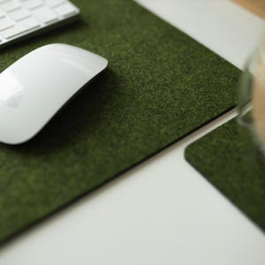 May include: A white wireless mouse sits on a green felt desk mat. The mat is textured to resemble grass. A glass of coffee sits on a matching green felt coaster.