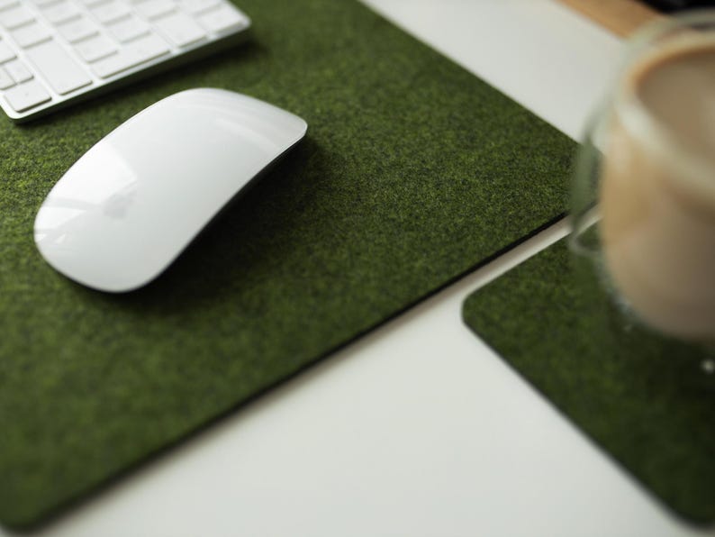 May include: A white wireless mouse sits on a green felt desk mat. The mat is textured to resemble grass. A glass of coffee sits on a matching green felt coaster.