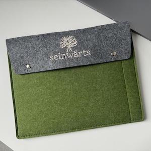 May include: A rectangular felt laptop sleeve featuring a green main body and a dark gray fold-over flap. The flap is secured with two silver metal snaps and displays an embroidered white tree icon above the text "seinwärts" in white thread.