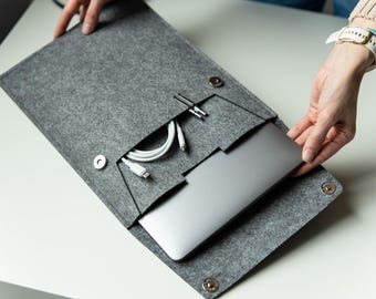 May include: A dark gray felt laptop sleeve featuring a dedicated front pocket for accessories. The sleeve includes a white charging cable stored in the pocket and a metallic snap button closure. A silver laptop is partially inserted into the main compartment.