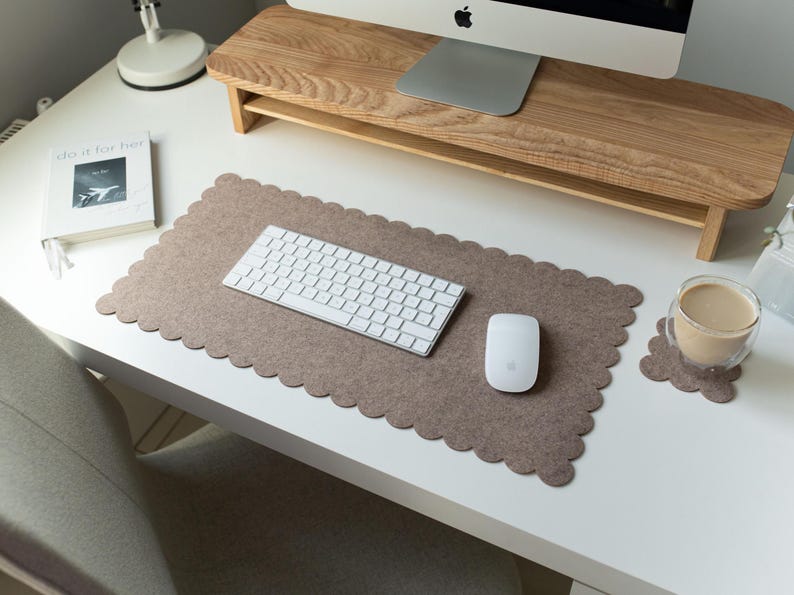 Puede incluir: Una moderna configuraci&oacute;n de escritorio con un soporte de monitor de madera, un teclado blanco y un rat&oacute;n blanco sobre una alfombrilla de escritorio marr&oacute;n con borde festoneado. Una taza de caf&eacute; descansa sobre un posavasos a juego. Tambi&eacute;n hay un libro con el texto "do it for her".