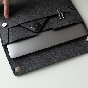 Op de afbeelding: Een grijze laptophuls van vilt met een ritssluiting en een klein vakje voor een telefoon of andere accessoires. De hoes is open en een laptop en telefoon zijn zichtbaar in de hoes.