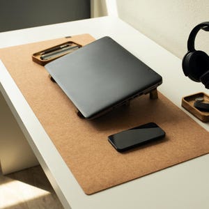 May include: A brown cork desk mat with a laptop, a smartphone, and a wooden headphone stand with black headphones on a white desk.