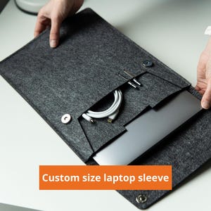 May include: A dark grey felt laptop sleeve with a flap closure secured by two silver-toned metal magnetic snaps. The sleeve features an integrated front pocket containing a white charging cable. The text "Custom size laptop sleeve" is visible.