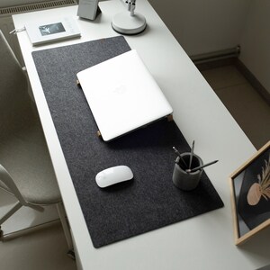 May include: A white laptop computer is open on a gray felt desk mat. A white wireless mouse is on the mat. A small gray concrete pencil holder with pencils is on the mat. A framed picture with a dried plant is on a white desk.