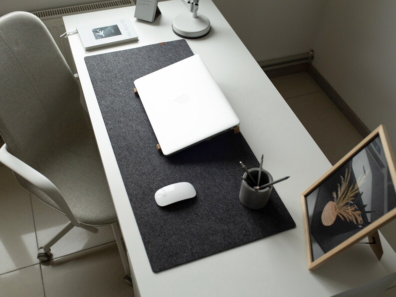 May include: A white laptop computer is open on a gray felt desk mat. A white wireless mouse is on the mat. A small gray concrete pencil holder with pencils is on the mat. A framed picture with a dried plant is on a white desk.