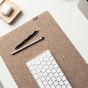 Pode incluir: Um tapete de secretária retangular de feltro castanho-claro com uma pequena etiqueta de tecido cinzento. Um teclado sem fios branco, uma esferográfica preta e um lápis de grafite cinzento estão sobre o tapete. Uma caixa de auriculares branca está num tabuleiro de madeira.