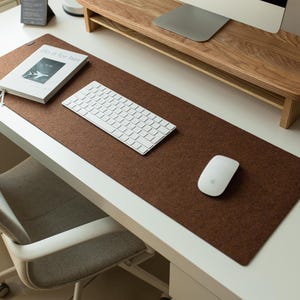 May include: A brown felt desk mat with a white keyboard and mouse on top. The desk mat is on a white desk with a wooden monitor stand.