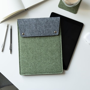 May include: A rectangular felt sleeve for a tablet, featuring a sage green body and a dark gray fold-over flap secured by two silver-toned metal snap buttons. The sleeve has a structured, flat design with visible stitching along the edges.