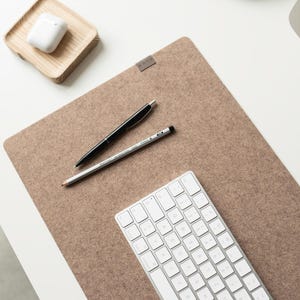 May include: A rectangular tan felt desk mat featuring a small grey fabric tag. A white wireless keyboard, a black ballpoint pen, and a grey graphite pencil are placed on the mat. A small wooden tray with white wireless earbuds sits nearby.