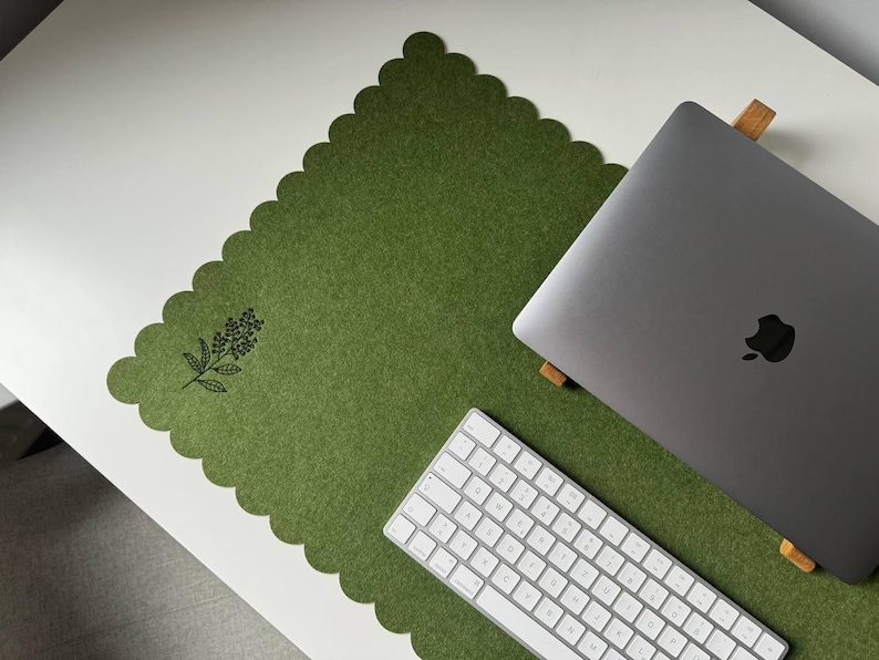 Puede incluir: Una alfombrilla de escritorio de fieltro verde con borde festoneado y dise&ntilde;o floral. Un port&aacute;til plateado, un teclado blanco y un soporte de madera para port&aacute;til est&aacute;n sobre la alfombrilla, que se encuentra sobre una superficie blanca.
