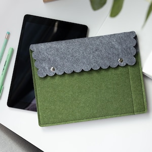 May include: A rectangular olive green felt sleeve with a scalloped grey felt flap secured by two silver metal snaps. The sleeve is positioned next to a black tablet and two light mint green pens, showcasing a structured design for device storage.