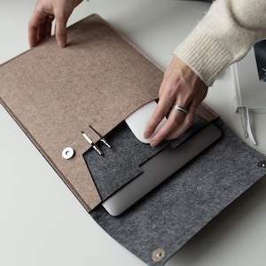 May include: A felt laptop sleeve in tan and gray, with a magnetic closure. The sleeve holds a laptop, a white mouse, and two silver pens. The sleeve is open, revealing the contents. A book with the text "for her" is in the background.